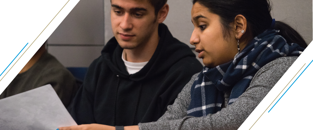 photo: two students sitting in lecture lean in close together as they confer over a piece of paper