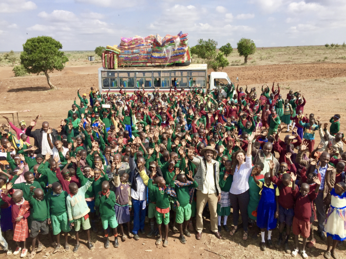 photo: Taking place in Kenya, GW student Francesca Diggs stands in the middle of a group of over 50 children in front of a school bus. The children, who are waving and smiling with excitement, are wearing vibrant green and red traditional uniforms. Behind them is the serene, vast landscape of Kenya
