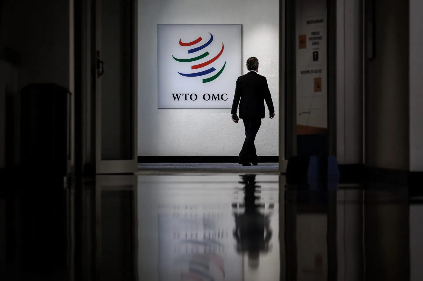 A man walking through the halls of the World Trade Organization. His back is facing the camera and he is walking by a sign that has their logo on the wall.
