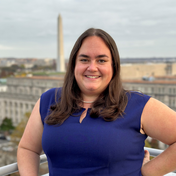 Erin Heffernan smiles. She is wearing a blue dress and pearl necklace. She is outside of the Elliott School City View Room overlooking the Washington Monument.