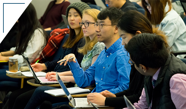 Photo: 5 students in a lecture hall, the student in the middle appears to be speaking to the professor