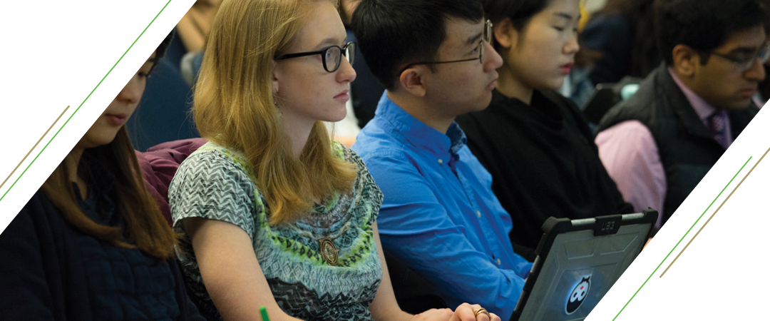 photo: four students sit attentively in a lecture hall, listening and watching an facing an unpictured lecturer that is out of the picture frame. One of the student's pictured, a girl with blonde hair and glasses, listens while taking notes on her laptop