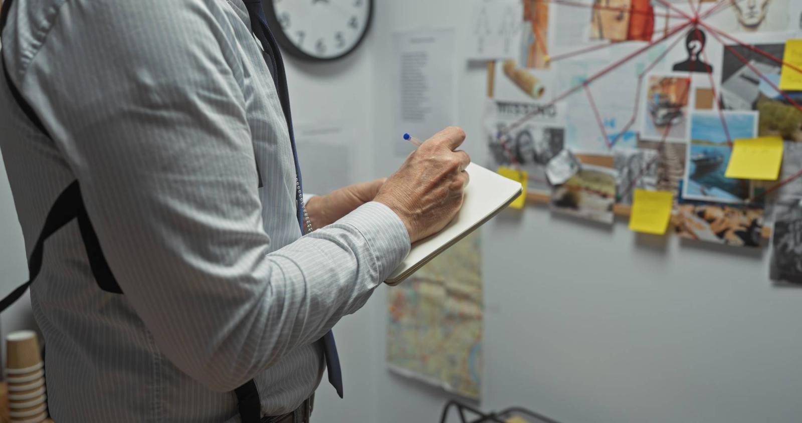 Man takes notes in a police station in front of a board connecting leads on a crime.